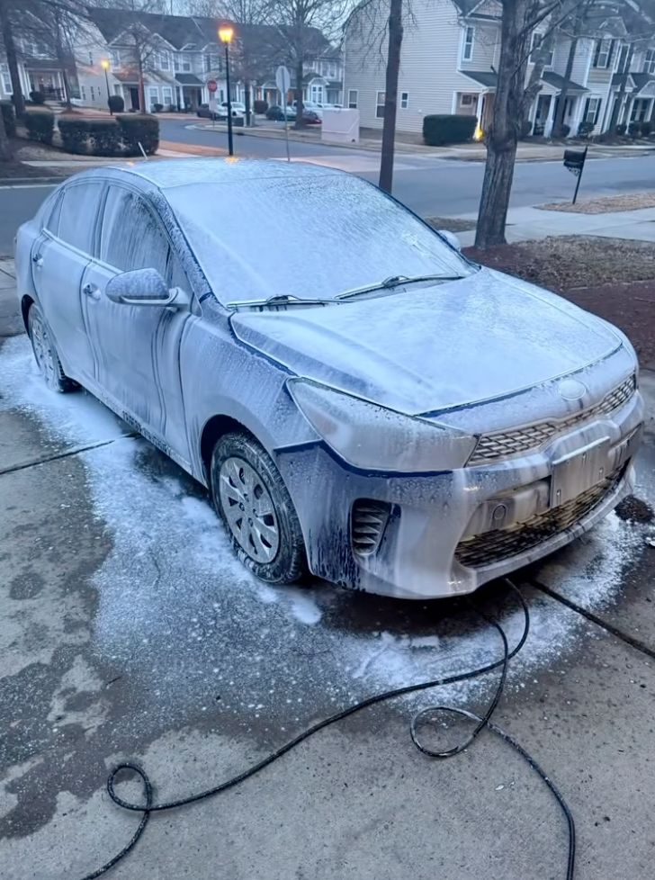 Silver sedan covered in ice on suburban street with garden hose nearby